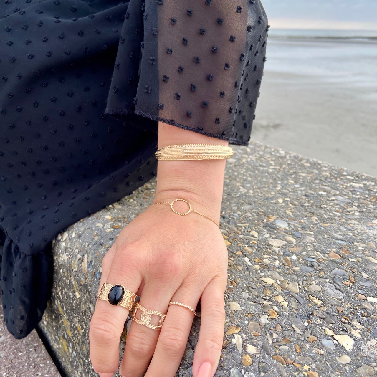 Main chic portant bagues et bracelets dorés élégants sur une plage, robe noire à pois