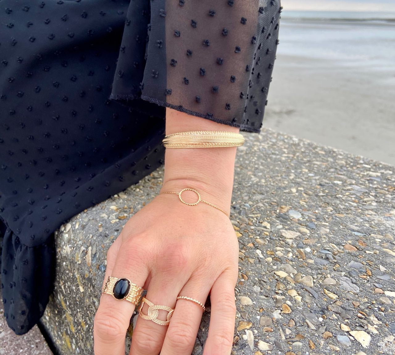 Main chic portant bagues et bracelets dorés élégants sur une plage, robe noire à pois