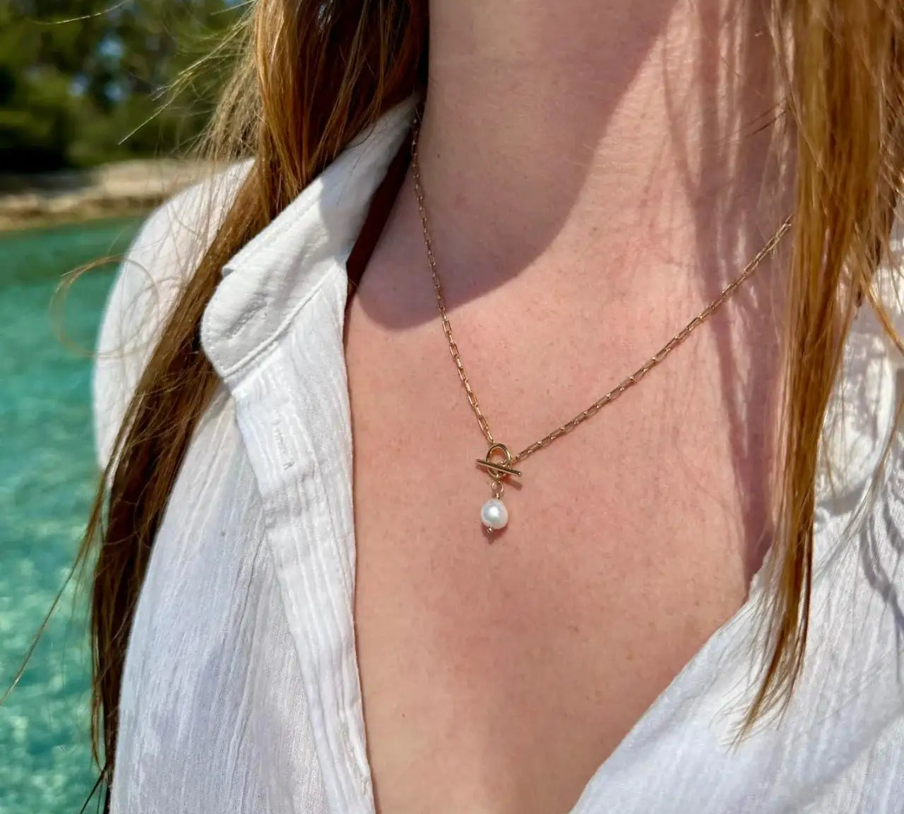 Collier doré avec perle, porté sur une chemise blanche au bord de l’eau