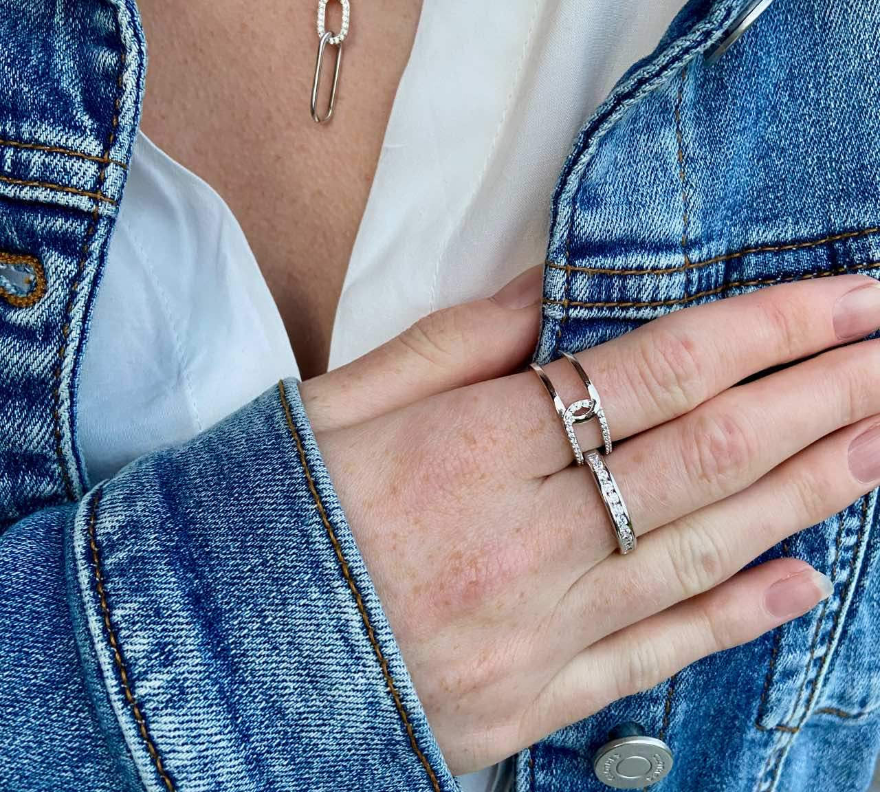Main portant deux bagues en argent rhodié et un collier épingle sur une veste en jean.
