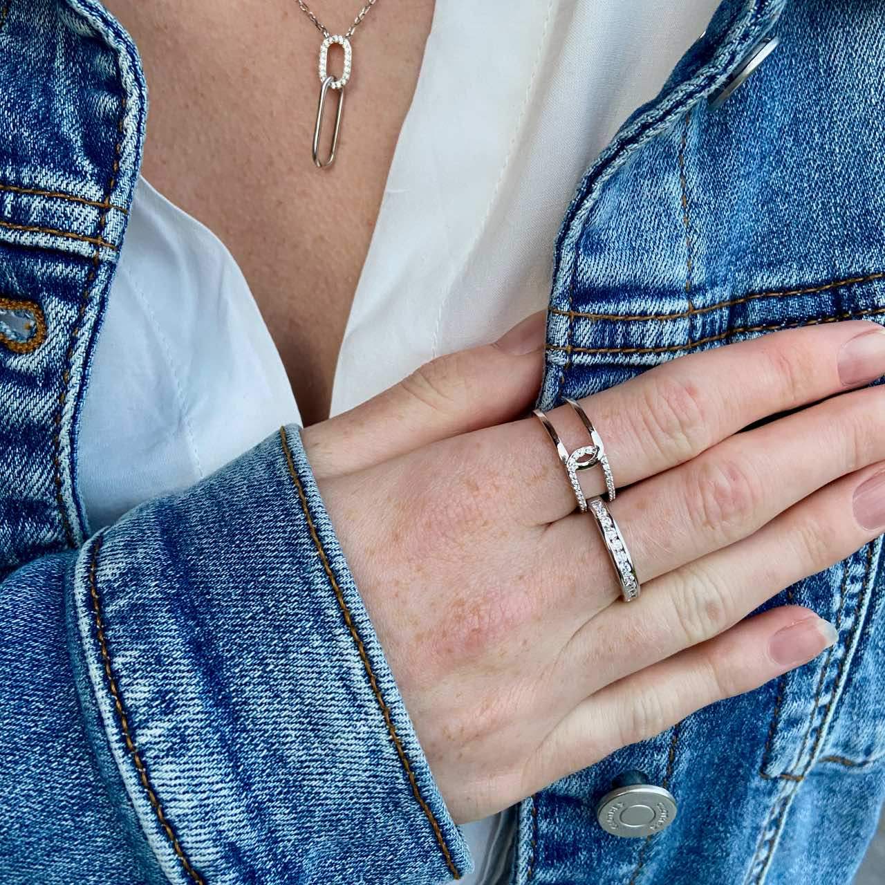 Main portant deux bagues en argent rhodié et un collier épingle sur une veste en jean.