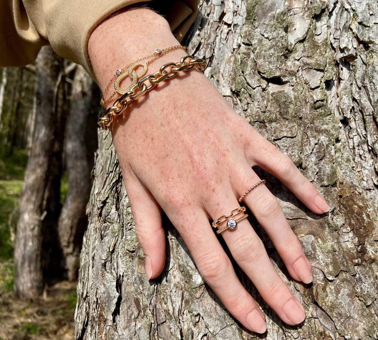 Main portant des bagues et bracelets en plaqué or posée sur l'écorce d'un arbre.
