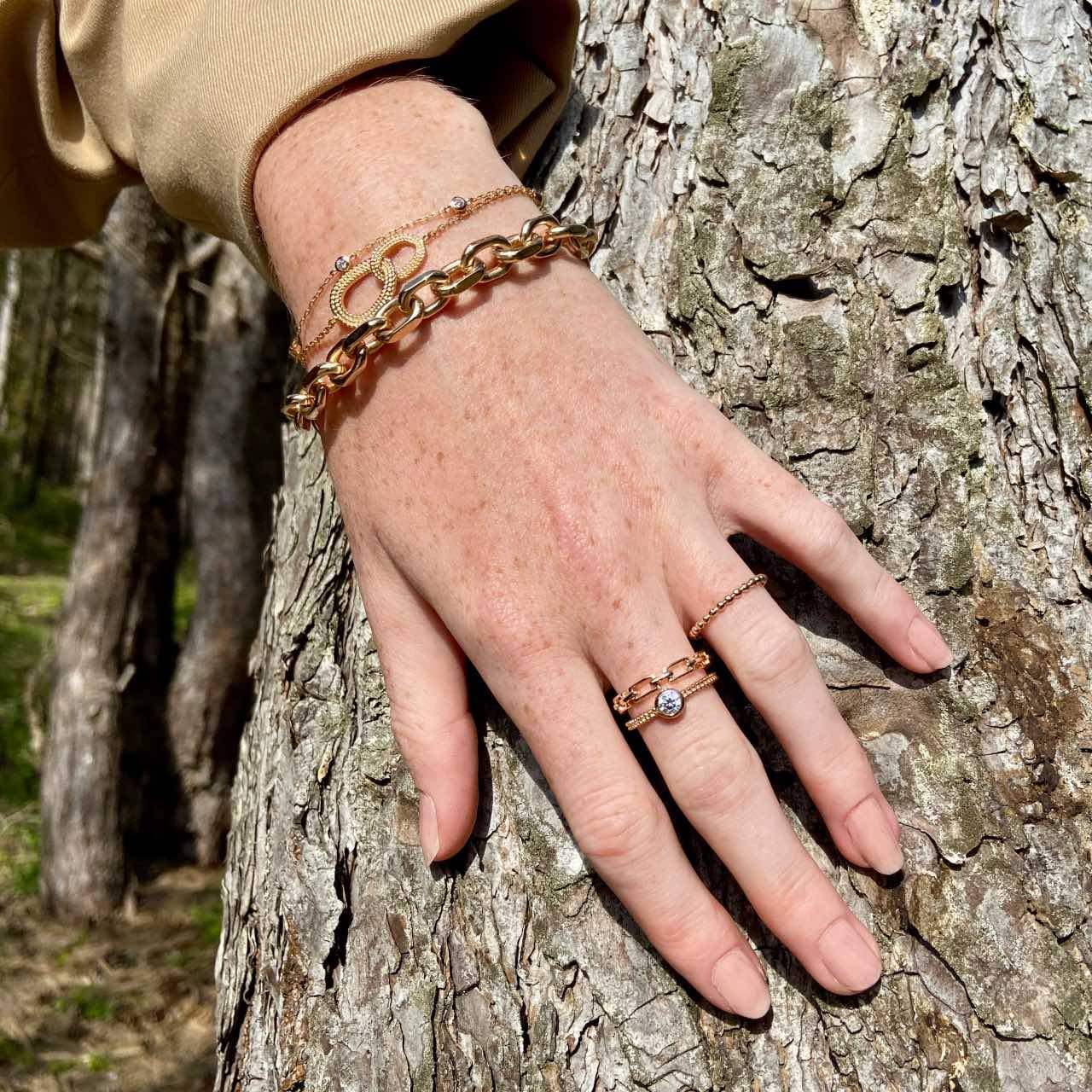 Main portant des bagues et bracelets en plaqué or posée sur l'écorce d'un arbre.